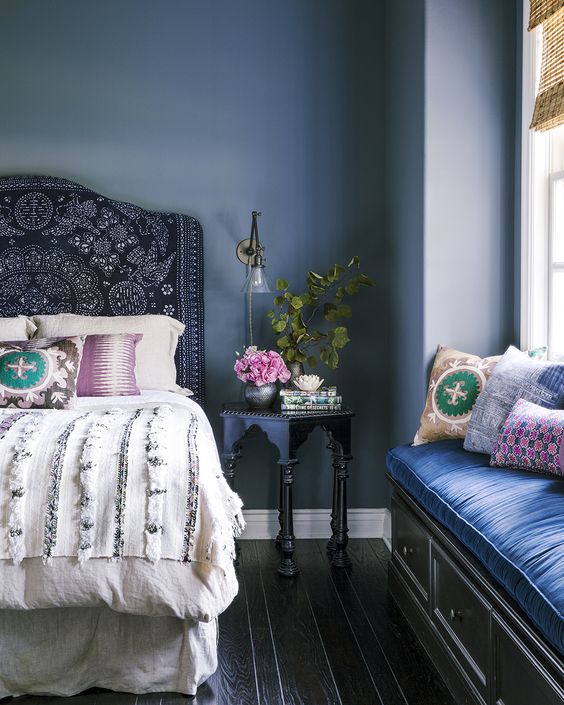 Bedroom designed by Amber Lewis. Blue fabric covered headboard with a white comforter with silver sequens. Decorative pillows on the bed. Dark wood floor with a blue wall. Morrocan style side table with flowers. Window seat with a blue cushion and colorful pillows